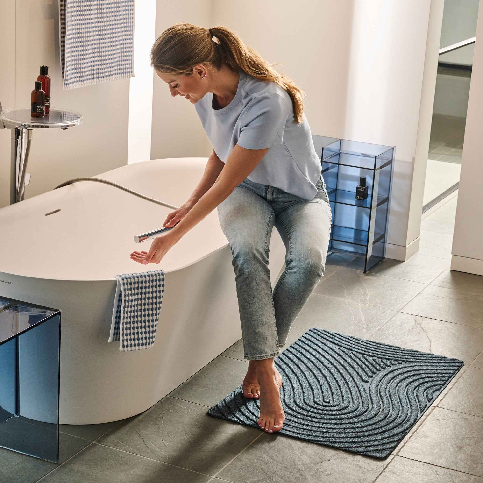 A woman tests bathwater while sitting on a freestanding tub, her feet on a textured blue bath mat in a modern bathroom.
