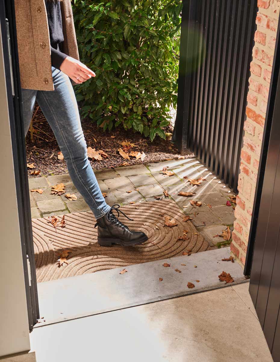 Outdoor entrance featuring a durable WaterHog doormat Fingerprint with a modern swirl design, capturing fallen autumn leaves as someone walks through the doorway.