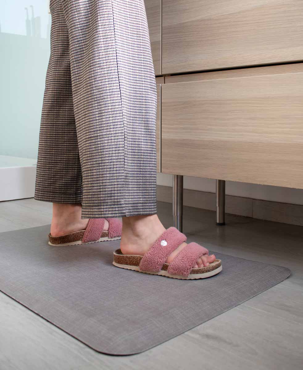 Grey Happy Feet mat in a modern bathroom, providing cushioned support as a person in pink fuzzy slippers stands near a wooden vanity.