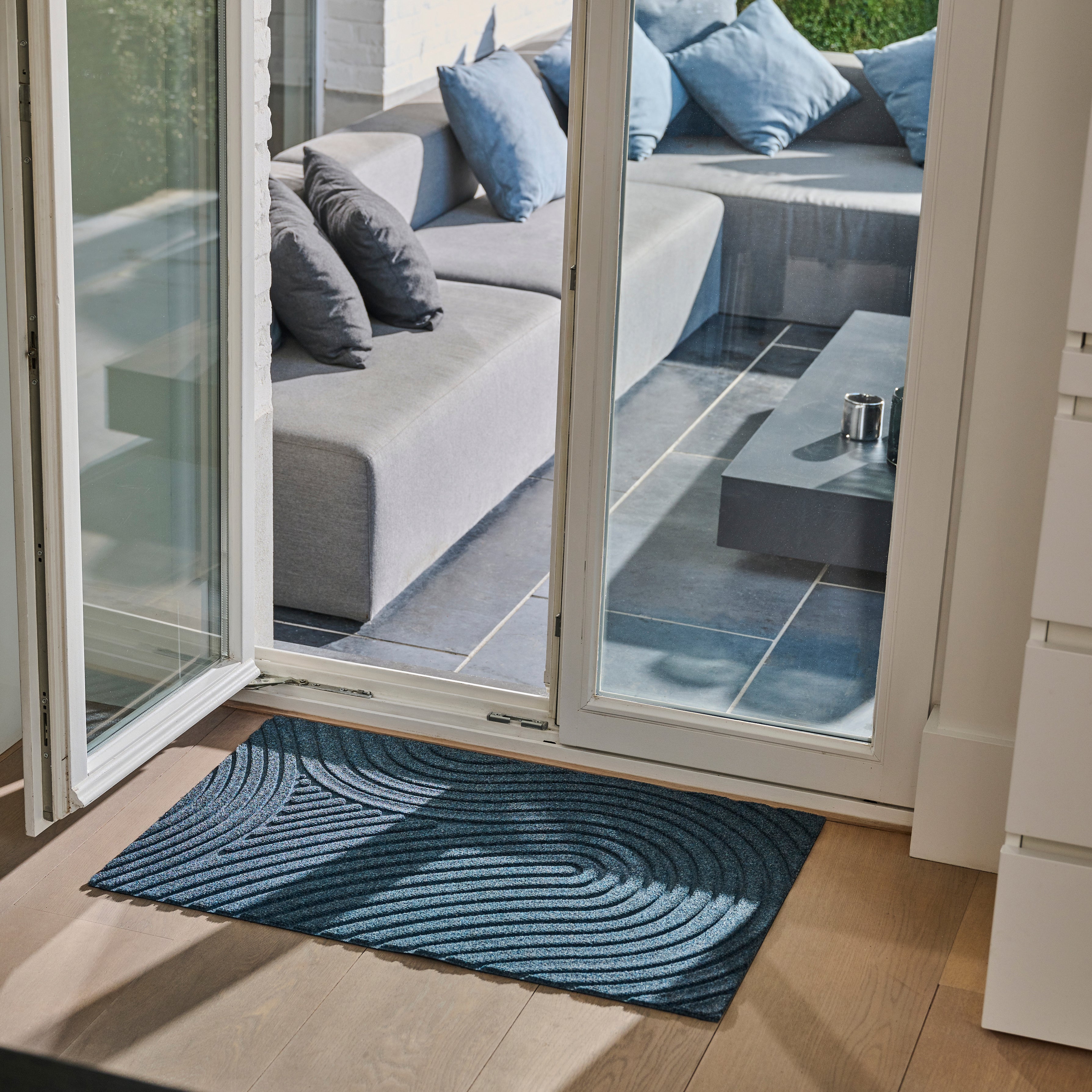 Lifestyle photo showcasing WaterHog All-Weather Indoor Outdoor blue navy marine colour Floor Mat in use in front of door at a terrace with a seat and pillows and an open door