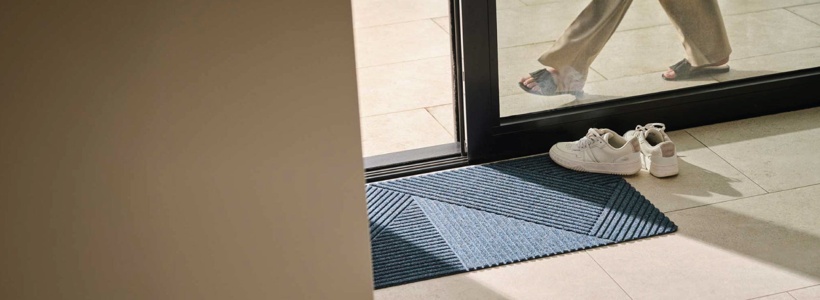 Stylish blue geometric doormat from the Matterly X WaterHog Collection placed at a glass door, with white sneakers beside it, enhancing a clean entryway.